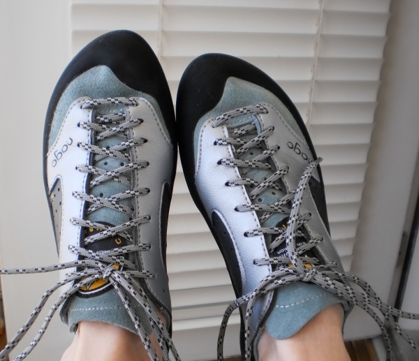 Two feet wearing new-looking rock climbing slippers. The shoes take up most of the frame with only two white ankles being visible. The shoes are silver and blue with a La Sportiva logo hidden under the shoelaces.